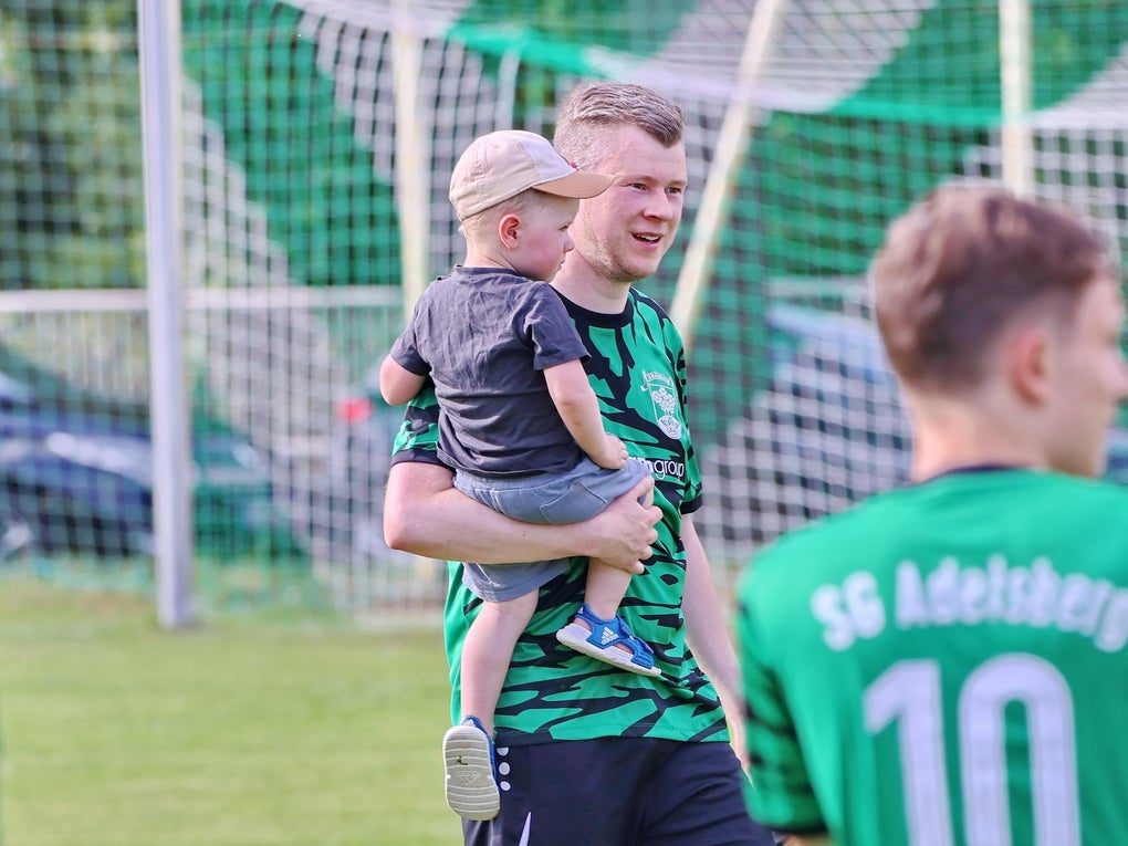 ein Mann steht auf einem Rasen, hinter ihm ein Fußballtor und er hält ein Kind auf dem Arm ein Mann steht auf einem Rasen, hinter ihm ein Fußballtor und er hält ein Kind auf dem Arm