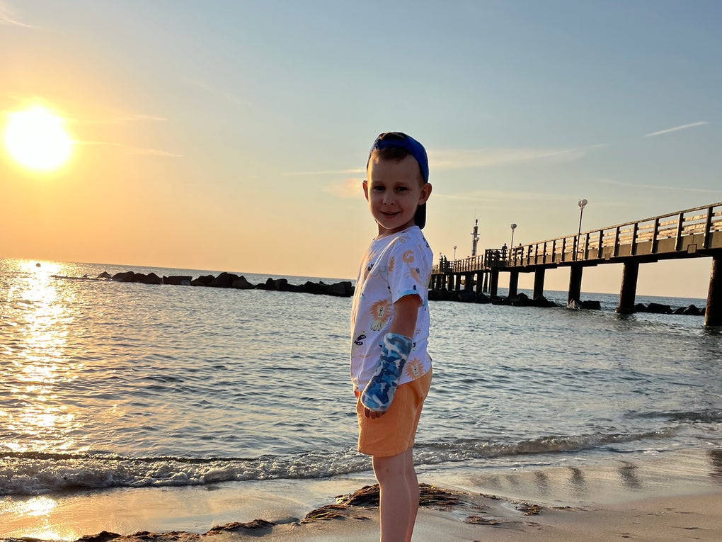 Ein Kind steht am Strand. Im Hintergrund sieht man das Meer, die untergehende Sonne und einen Steg Ein Kind steht am Strand. Im Hintergrund sieht man das Meer, die untergehende Sonne und einen Steg
