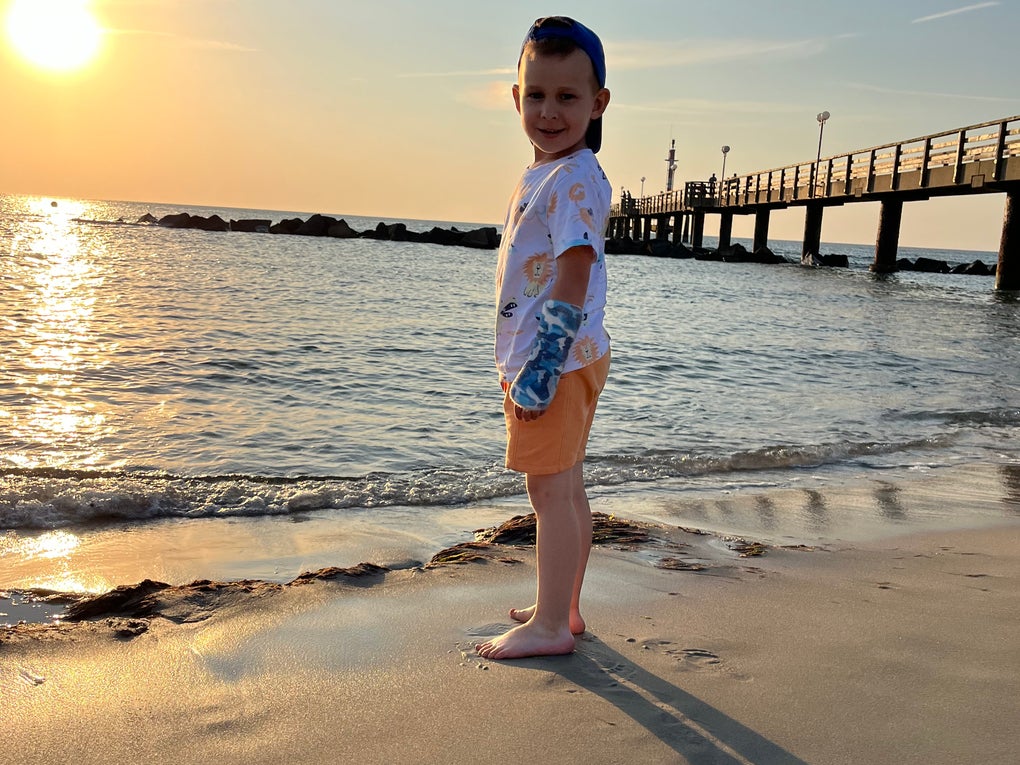 Ein Kind steht am Strand. Im Hintergrund sieht man das Meer, die untergehende Sonne und einen Steg Ein Kind steht am Strand. Im Hintergrund sieht man das Meer, die untergehende Sonne und einen Steg
