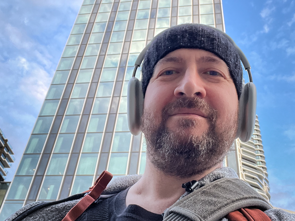 Ein Mann mit Kopfhörern und einer Mütze steht vor einem Hochhaus Ein Mann mit Kopfhörern und einer Mütze steht vor einem Hochhaus