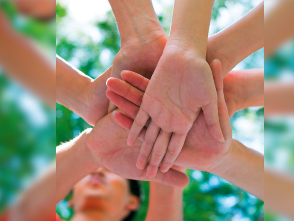 Hände werden in die Mitte gehalten und aufeinander gelegt. Im Hintergrund ist verschwommen ein Gesicht und ein Blätterdach erkennbar. Hände werden in die Mitte gehalten und aufeinander gelegt. Im Hintergrund ist verschwommen ein Gesicht und ein Blätterdach erkennbar.