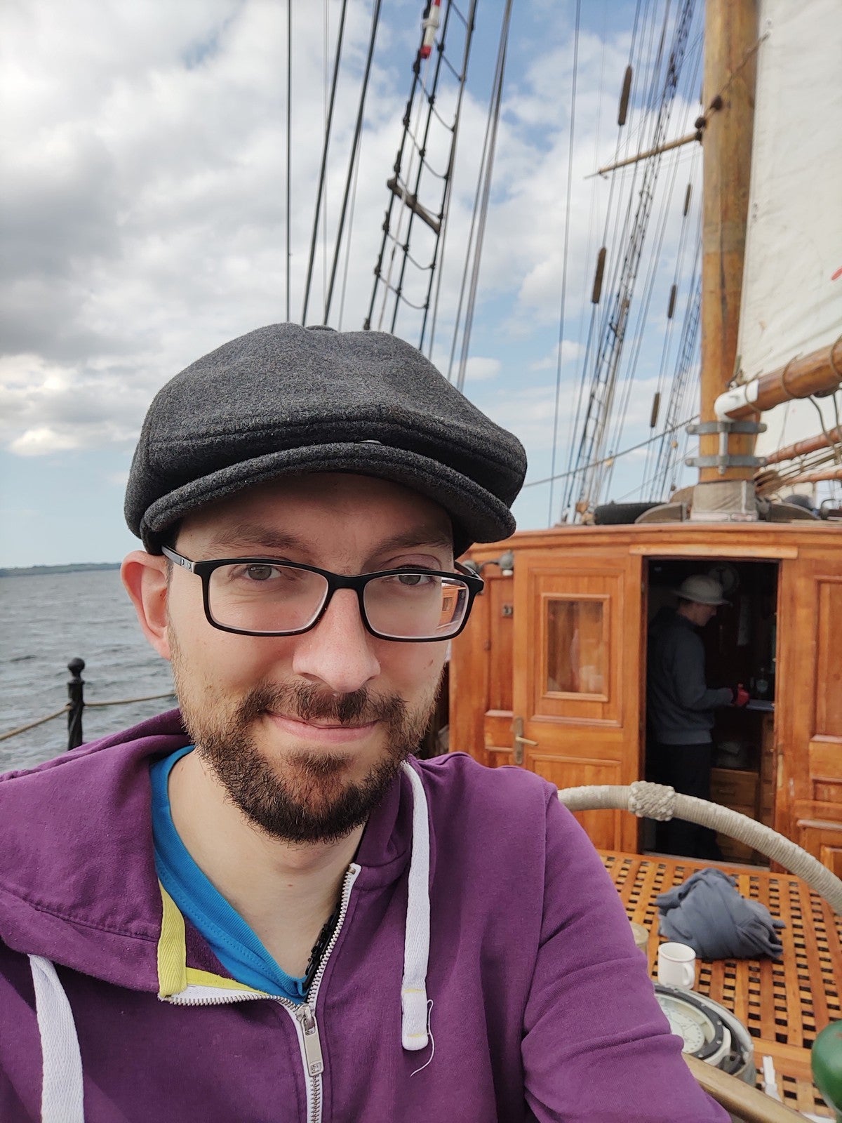 Ein Mann mit Brille, Bart und Schiebermütze trägt eine lila Kapuzenjacke und macht ein Selfie auf einem Segelschiff. Im Hintergrund sind ein hölzerner Aufbau mit geöffneter Tür, Seile, Masten und das Meer unter einem teilweise bewölkten Himmel zu sehen.
Ein Mann mit Brille, Bart und Schiebermütze trägt eine lila Kapuzenjacke und macht ein Selfie auf einem Segelschiff. Im Hintergrund sind ein hölzerner Aufbau mit geöffneter Tür, Seile, Masten und das Meer unter einem teilweise bewölkten Himmel zu sehen.