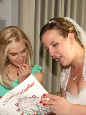 Eine Frau in einem Hochzeitskleid schaut in ein Buch, daneben steht eine andere Frau die mit in das Buch schaut
