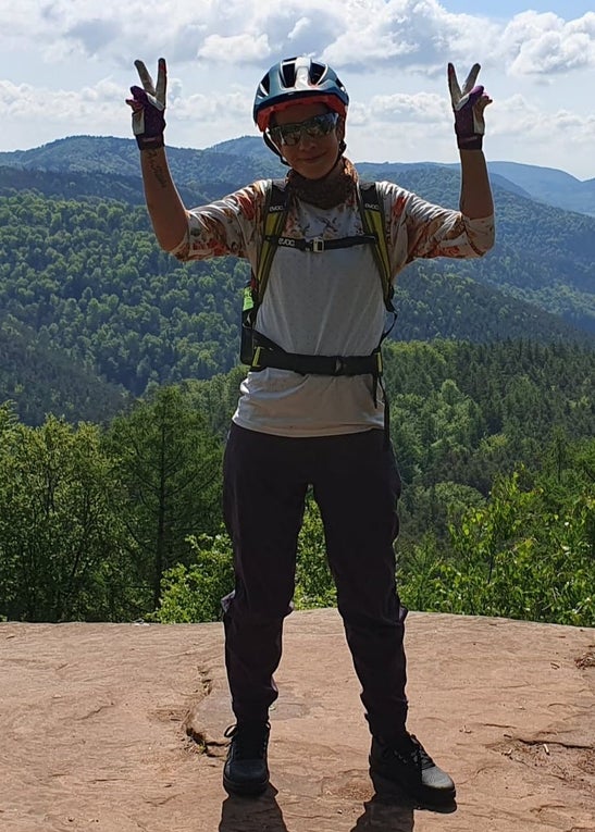 Ilona steht in Fahrradkleidung vor einer Bergkulisse. Sie lächelt in die Kamera und hält beide Hände zu Peace-Zeichen hoch. Ilona steht in Fahrradkleidung vor einer Bergkulisse. Sie lächelt in die Kamera und hält beide Hände zu Peace-Zeichen hoch.