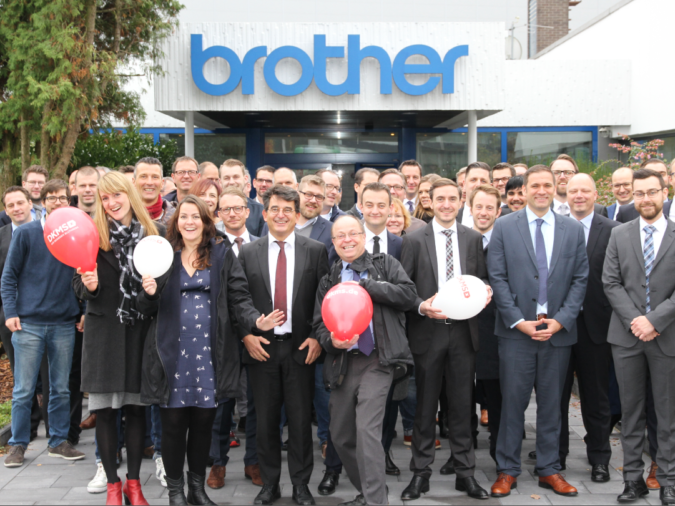 Eine Gruppe von Menschen posiert vor einem Gebäude. Auf dem Gebäude ist ein Logo der Firma brother. Eine Gruppe von Menschen posiert vor einem Gebäude. Auf dem Gebäude ist ein Logo der Firma brother.