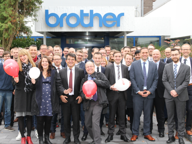 Eine Gruppe von Menschen posiert vor einem Gebäude. Auf dem Gebäude ist ein Logo der Firma brother. Eine Gruppe von Menschen posiert vor einem Gebäude. Auf dem Gebäude ist ein Logo der Firma brother.
