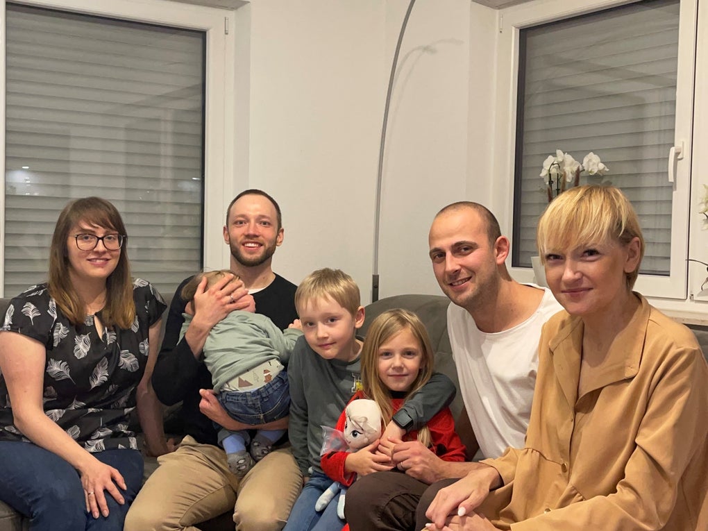 Gruppenfoto in einem Wohnzimmer: Sieben Personen sitzen eng beieinander auf einem Sofa und einem Hocker. Zu sehen sind drei Erwachsene, zwei Männer mit kurzen Haaren, eine Frau mit blonder Kurzhaarfrisur, daneben eine weitere Frau mit Brille, ein Baby auf dem Arm eines Mannes sowie zwei Kinder in der Mitte, ein Junge und ein Mädchen mit Kuscheltier. Alle lächeln in die Kamera. Gruppenfoto in einem Wohnzimmer: Sieben Personen sitzen eng beieinander auf einem Sofa und einem Hocker. Zu sehen sind drei Erwachsene, zwei Männer mit kurzen Haaren, eine Frau mit blonder Kurzhaarfrisur, daneben eine weitere Frau mit Brille, ein Baby auf dem Arm eines Mannes sowie zwei Kinder in der Mitte, ein Junge und ein Mädchen mit Kuscheltier. Alle lächeln in die Kamera.