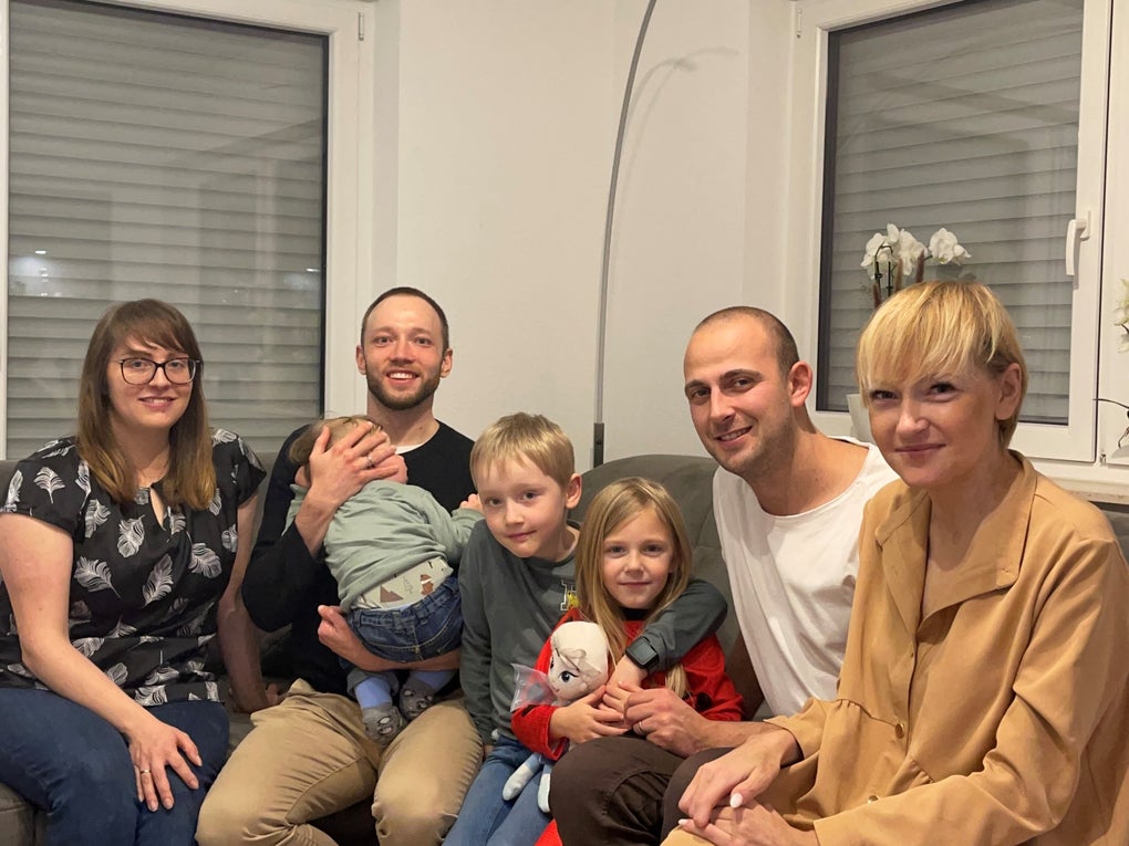 Gruppenfoto in einem Wohnzimmer: Sieben Personen sitzen eng beieinander auf einem Sofa und einem Hocker. Zu sehen sind drei Erwachsene, zwei Männer mit kurzen Haaren, eine Frau mit blonder Kurzhaarfrisur, daneben eine weitere Frau mit Brille, ein Baby auf dem Arm eines Mannes sowie zwei Kinder in der Mitte, ein Junge und ein Mädchen mit Kuscheltier. Alle lächeln in die Kamera. Gruppenfoto in einem Wohnzimmer: Sieben Personen sitzen eng beieinander auf einem Sofa und einem Hocker. Zu sehen sind drei Erwachsene, zwei Männer mit kurzen Haaren, eine Frau mit blonder Kurzhaarfrisur, daneben eine weitere Frau mit Brille, ein Baby auf dem Arm eines Mannes sowie zwei Kinder in der Mitte, ein Junge und ein Mädchen mit Kuscheltier. Alle lächeln in die Kamera.