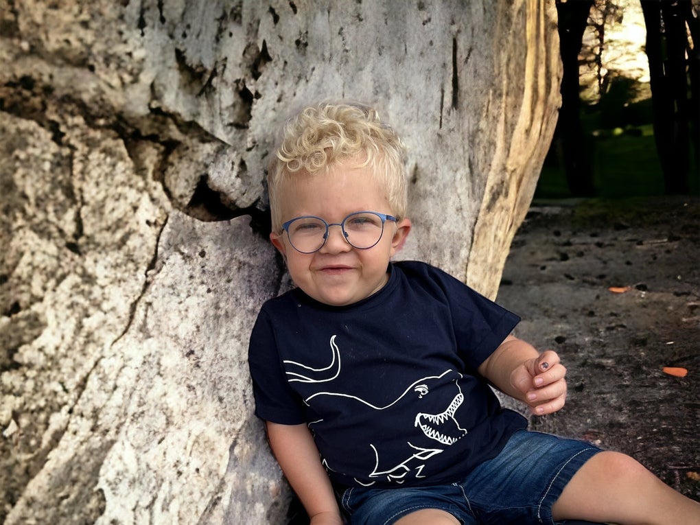 kleines Kind mit blonden Haaren und Brille sitzt auf dem Boden und lehnt an einen Baum kleines Kind mit blonden Haaren und Brille sitzt auf dem Boden und lehnt an einen Baum