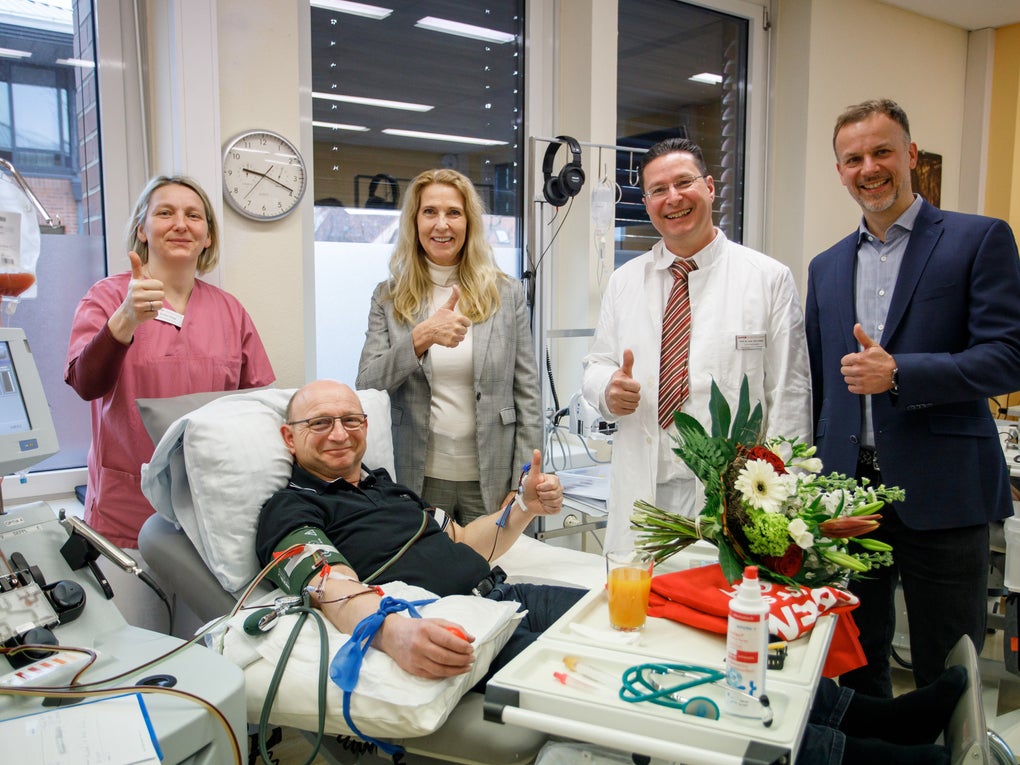 Ein Mann liegt in einem Spendenbett und spendet Blutstammzellen. Er ist an ein Apherese-Gerät angeschlossen. Neben ihm stehen vier Personen, zwei Männer im Anzug bzw. Kittel, eine Frau in medizinischer Kleidung und eine Frau im Hosenanzug. Alle fünf Personen zeigen den Daumen nach oben. Auf dem Tisch vor dem Spender stehen ein Blumenstrauß, ein Glas Orangensaft und rote Stoffe. Im Hintergrund sind medizinische Geräte, Fenster, Uhren und eine weiße Wand zu sehen.
Ein Mann liegt in einem Spendenbett und spendet Blutstammzellen. Er ist an ein Apherese-Gerät angeschlossen. Neben ihm stehen vier Personen, zwei Männer im Anzug bzw. Kittel, eine Frau in medizinischer Kleidung und eine Frau im Hosenanzug. Alle fünf Personen zeigen den Daumen nach oben. Auf dem Tisch vor dem Spender stehen ein Blumenstrauß, ein Glas Orangensaft und rote Stoffe. Im Hintergrund sind medizinische Geräte, Fenster, Uhren und eine weiße Wand zu sehen.