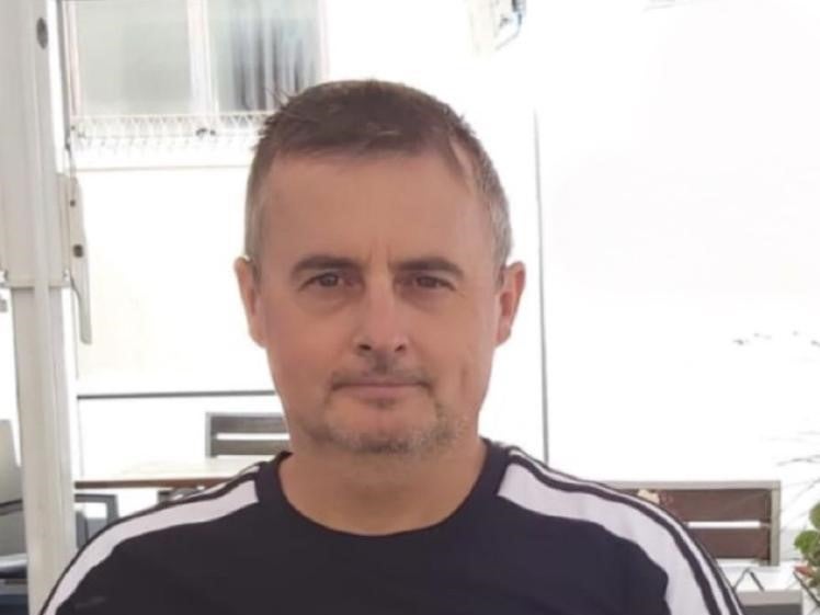 Ein Mann mit kurzem Haar und leichtem Bart sitzt draußen vor einem hellen Hintergrund. Er trägt ein dunkelblaues oder schwarzes T-Shirt mit weißen Streifen an den Schultern. Im Hintergrund sind Fenster, ein Tisch und helle Strukturen zu sehen. Ein Mann mit kurzem Haar und leichtem Bart sitzt draußen vor einem hellen Hintergrund. Er trägt ein dunkelblaues oder schwarzes T-Shirt mit weißen Streifen an den Schultern. Im Hintergrund sind Fenster, ein Tisch und helle Strukturen zu sehen.