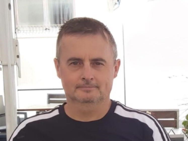 Ein Mann mit kurzem Haar und leichtem Bart sitzt draußen vor einem hellen Hintergrund. Er trägt ein dunkelblaues oder schwarzes T-Shirt mit weißen Streifen an den Schultern. Im Hintergrund sind Fenster, ein Tisch und helle Strukturen zu sehen. Ein Mann mit kurzem Haar und leichtem Bart sitzt draußen vor einem hellen Hintergrund. Er trägt ein dunkelblaues oder schwarzes T-Shirt mit weißen Streifen an den Schultern. Im Hintergrund sind Fenster, ein Tisch und helle Strukturen zu sehen.