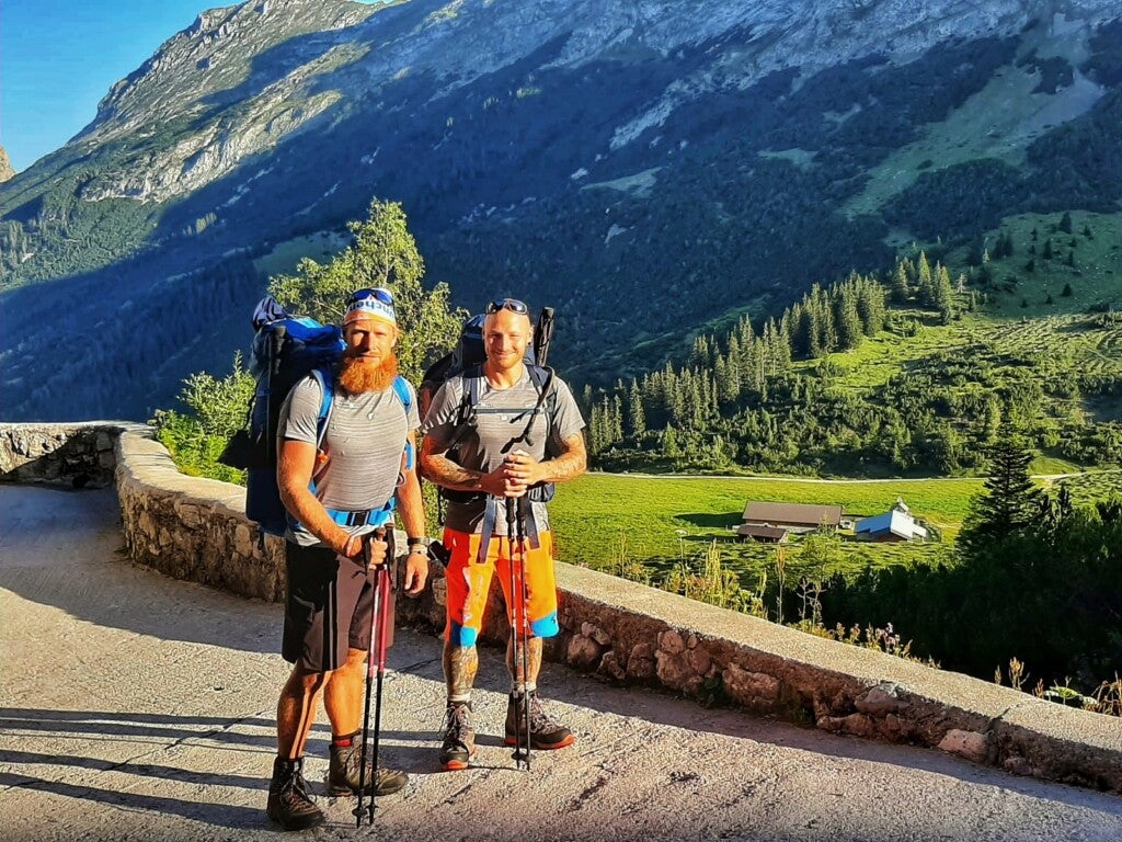 Zwei Männer mit Rucksäcken stehen auf einer Straße mit einem Berg im Hintergrund Zwei Männer mit Rucksäcken stehen auf einer Straße mit einem Berg im Hintergrund