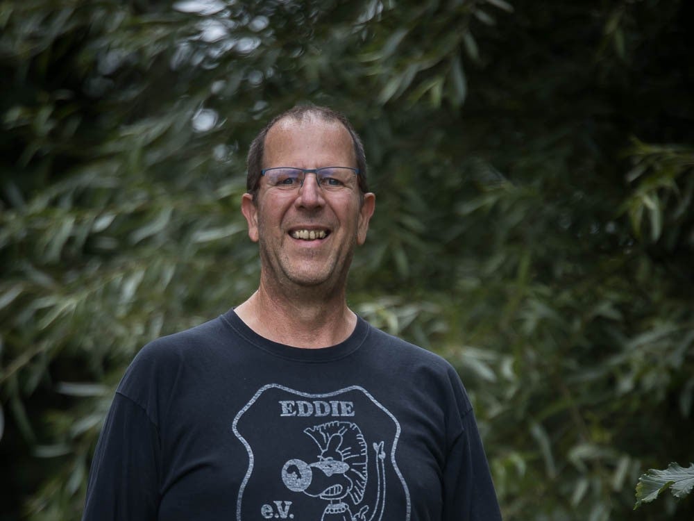 ein Mann mit schwarzem T-Shirt und Brille lacht in die Kamera. Im Hintergrund sieht man Bäume ein Mann mit schwarzem T-Shirt und Brille lacht in die Kamera. Im Hintergrund sieht man Bäume