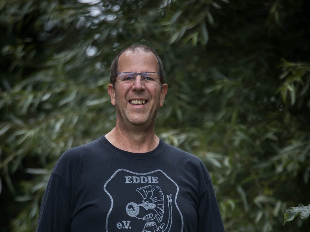 ein Mann mit schwarzem T-Shirt und Brille lacht in die Kamera. Im Hintergrund sieht man Bäume ein Mann mit schwarzem T-Shirt und Brille lacht in die Kamera. Im Hintergrund sieht man Bäume