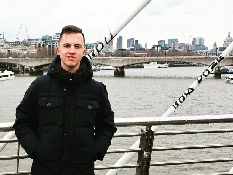 Ein Mann steht auf einer Brücke mit Metallgeländer vor einem Fluss. Er trägt eine schwarze Jacke und blaue Jeans. Im Hintergrund sind eine weitere Brücke, Boote auf dem Wasser und eine Stadtsilhouette mit modernen und historischen Gebäuden zu sehen. Der Himmel ist bewölkt.  Ein Mann steht auf einer Brücke mit Metallgeländer vor einem Fluss. Er trägt eine schwarze Jacke und blaue Jeans. Im Hintergrund sind eine weitere Brücke, Boote auf dem Wasser und eine Stadtsilhouette mit modernen und historischen Gebäuden zu sehen. Der Himmel ist bewölkt.