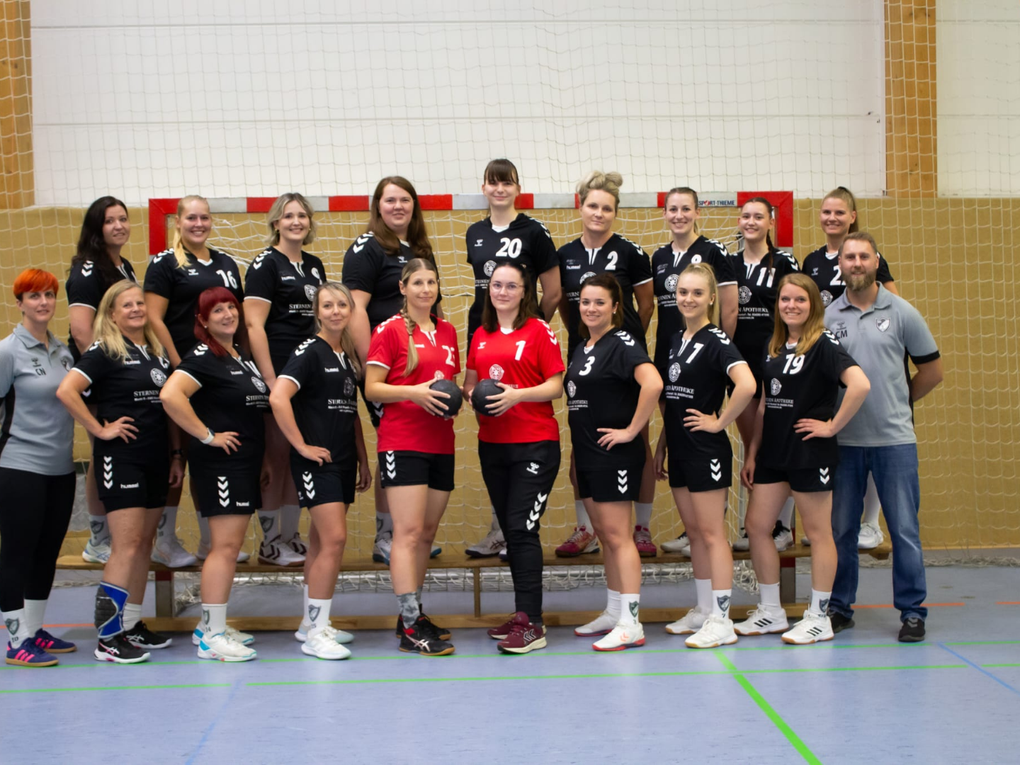 Eine Damenhandballmannschaft steht mit zwei Trainer:innen vor einem Tor. Eine Damenhandballmannschaft steht mit zwei Trainer:innen vor einem Tor.