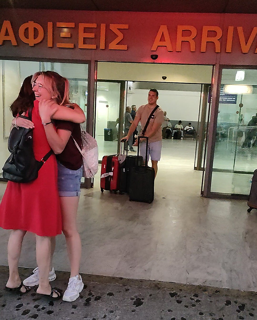 Vor dem Ausgangsbereich eines Flughafens mit der Aufschrift „Ankünfte“ auf Griechisch und Englisch umarmen sich zwei Frauen. Eine trägt ein rotes Kleid, die andere ein bordeauxrotes Oberteil und Jeansshorts. Im Hintergrund schiebt ein junger Mann einen Gepäckwagen mit mehreren Koffern durch die Glastür in ihre Richtung. Vor dem Ausgangsbereich eines Flughafens mit der Aufschrift „Ankünfte“ auf Griechisch und Englisch umarmen sich zwei Frauen. Eine trägt ein rotes Kleid, die andere ein bordeauxrotes Oberteil und Jeansshorts. Im Hintergrund schiebt ein junger Mann einen Gepäckwagen mit mehreren Koffern durch die Glastür in ihre Richtung.