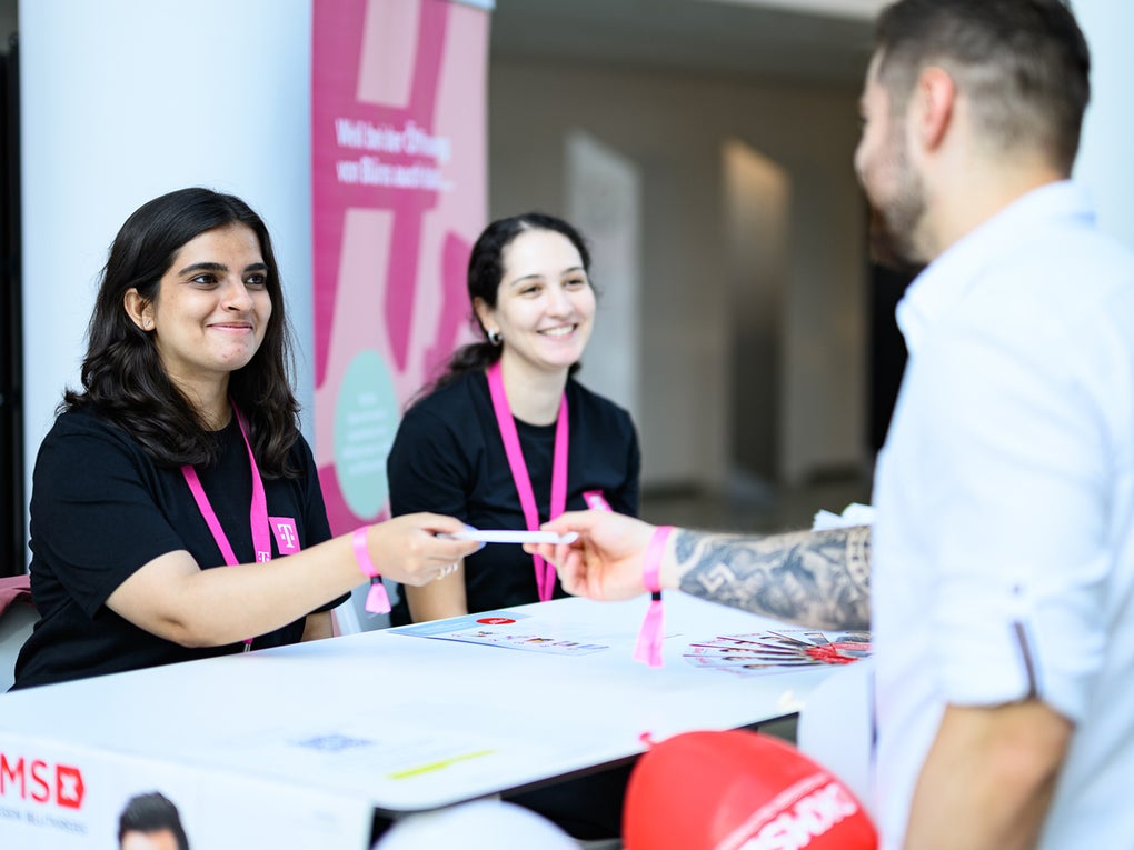 Zwei junge Frauen mit pinken DKMS-Lanyards sitzen hinter einem Infostand und lächeln. Eine von ihnen überreicht einem Mann im Vordergrund ein Formular oder Set. Der Mann steht mit dem Rücken zur Kamera. Auf dem Tisch sind DKMS-Logos, Flyer und rote Luftballons zu sehen. Im Hintergrund ist ein Banner mit dem Schriftzug „Herzlich willkommen“.
Zwei junge Frauen mit pinken DKMS-Lanyards sitzen hinter einem Infostand und lächeln. Eine von ihnen überreicht einem Mann im Vordergrund ein Formular oder Set. Der Mann steht mit dem Rücken zur Kamera. Auf dem Tisch sind DKMS-Logos, Flyer und rote Luftballons zu sehen. Im Hintergrund ist ein Banner mit dem Schriftzug „Herzlich willkommen“.