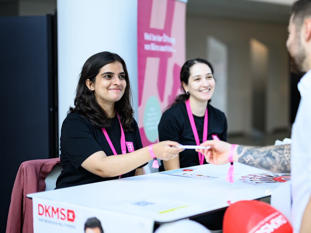 Zwei junge Frauen mit pinken DKMS-Lanyards sitzen hinter einem Infostand und lächeln. Eine von ihnen überreicht einem Mann im Vordergrund ein Formular oder Set. Der Mann steht mit dem Rücken zur Kamera. Auf dem Tisch sind DKMS-Logos, Flyer und rote Luftballons zu sehen. Im Hintergrund ist ein Banner mit dem Schriftzug „Herzlich willkommen“.
Zwei junge Frauen mit pinken DKMS-Lanyards sitzen hinter einem Infostand und lächeln. Eine von ihnen überreicht einem Mann im Vordergrund ein Formular oder Set. Der Mann steht mit dem Rücken zur Kamera. Auf dem Tisch sind DKMS-Logos, Flyer und rote Luftballons zu sehen. Im Hintergrund ist ein Banner mit dem Schriftzug „Herzlich willkommen“.