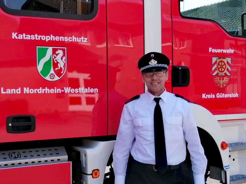 Ein Feuerwehrmann in weißem Hemd, schwarzer Krawatte und Uniformmütze steht vor einem roten Einsatzfahrzeug. Auf dem Fahrzeug steht „Katastrophenschutz Land Nordrhein-Westfalen“ sowie „Feuerwehr Kreis Gütersloh“, daneben sind die Landes- und Kreiswappen abgebildet. Ein Feuerwehrmann in weißem Hemd, schwarzer Krawatte und Uniformmütze steht vor einem roten Einsatzfahrzeug. Auf dem Fahrzeug steht „Katastrophenschutz Land Nordrhein-Westfalen“ sowie „Feuerwehr Kreis Gütersloh“, daneben sind die Landes- und Kreiswappen abgebildet.