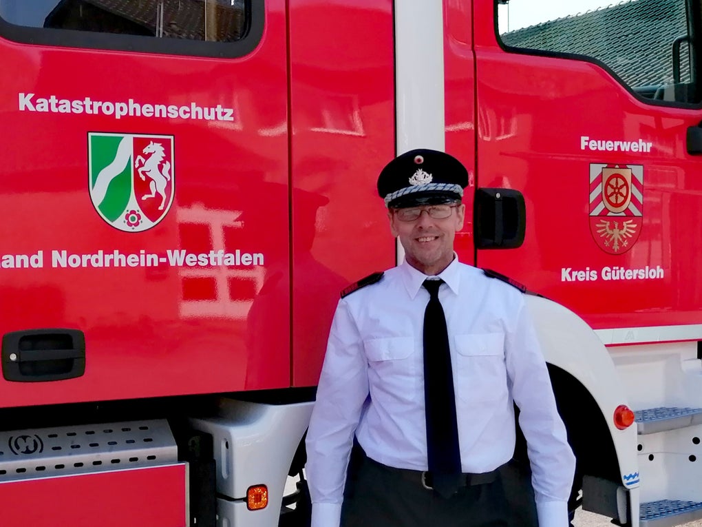 Ein Feuerwehrmann in weißem Hemd, schwarzer Krawatte und Uniformmütze steht vor einem roten Einsatzfahrzeug. Auf dem Fahrzeug steht „Katastrophenschutz Land Nordrhein-Westfalen“ sowie „Feuerwehr Kreis Gütersloh“, daneben sind die Landes- und Kreiswappen abgebildet. Ein Feuerwehrmann in weißem Hemd, schwarzer Krawatte und Uniformmütze steht vor einem roten Einsatzfahrzeug. Auf dem Fahrzeug steht „Katastrophenschutz Land Nordrhein-Westfalen“ sowie „Feuerwehr Kreis Gütersloh“, daneben sind die Landes- und Kreiswappen abgebildet.