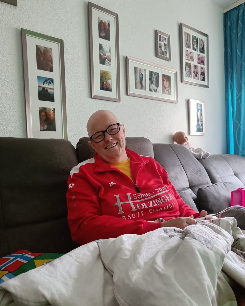 Patientin Manuela sitzt in ihrem Wohnzimmer vor einer Fotowand und lächelt