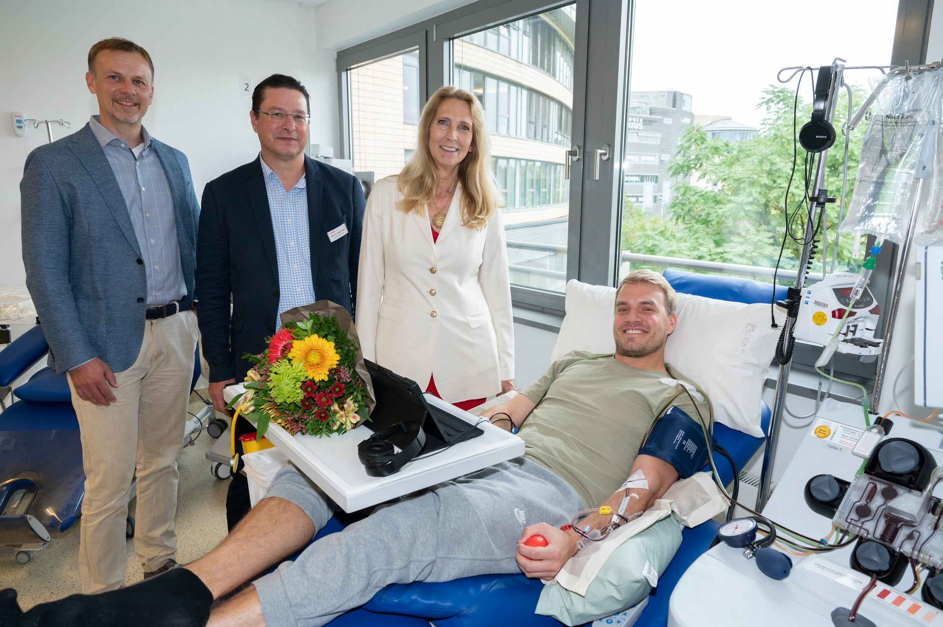 Eine Gruppe von Menschen steht um einen Mann in einem Krankenhausbett. Der Mann spendet gerade Stammzellen Eine Gruppe von Menschen steht um einen Mann in einem Krankenhausbett. Der Mann spendet gerade Stammzellen