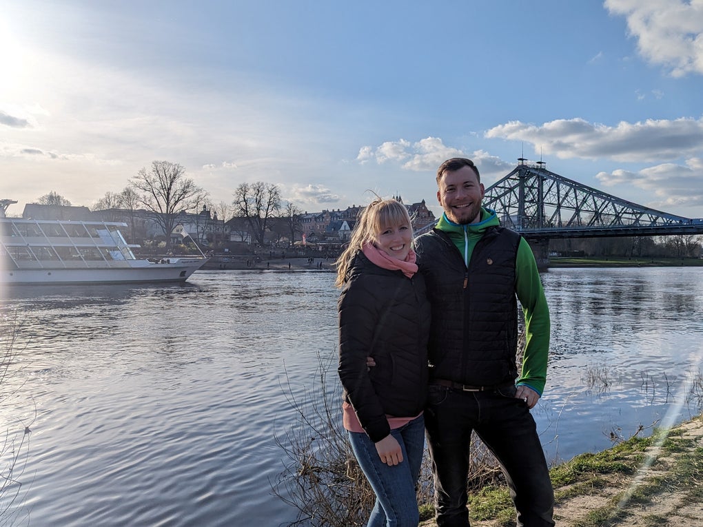 Eine Frau und ein Mann stehen an einem Flußufer, im Hintergrund sind ein Ausflugsschiff und eine Brücke zu sehen. Eine Frau und ein Mann stehen an einem Flußufer, im Hintergrund sind ein Ausflugsschiff und eine Brücke zu sehen.