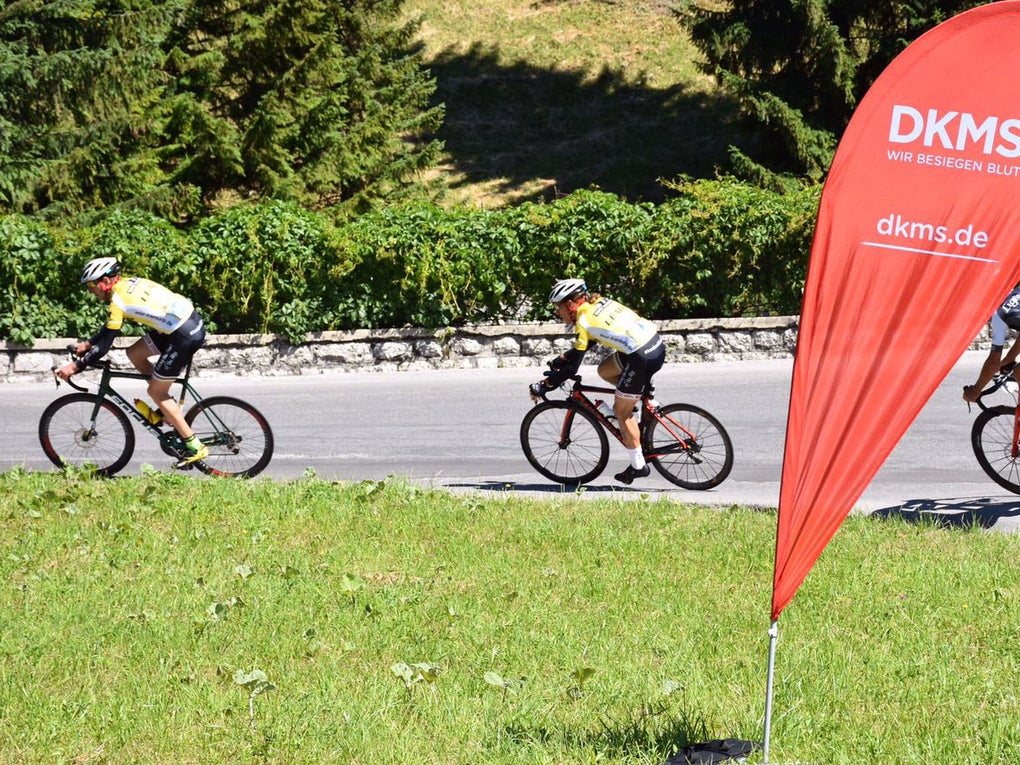 Eine Gruppe von Menschen auf Fahrrädern auf einer Straße. Im Vordergrund steht eine DKMS Flagge. Eine Gruppe von Menschen auf Fahrrädern auf einer Straße. Im Vordergrund steht eine DKMS Flagge.