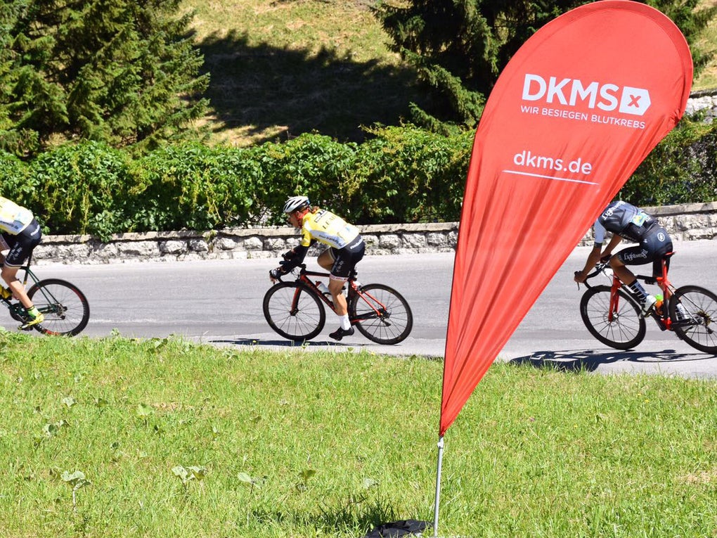 Eine Gruppe von Menschen auf Fahrrädern auf einer Straße. Im Vordergrund steht eine DKMS Flagge.  Eine Gruppe von Menschen auf Fahrrädern auf einer Straße. Im Vordergrund steht eine DKMS Flagge.