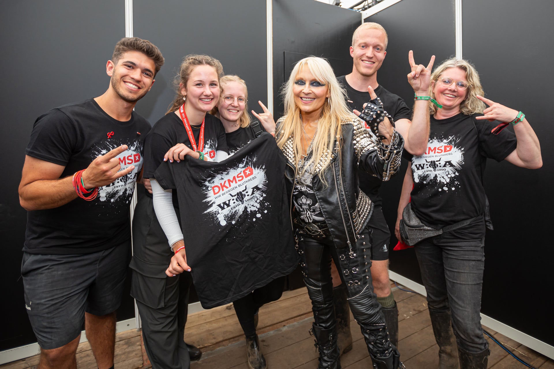Sechs Personen stehen vor einer dunklen Wand und posieren für ein Gruppenfoto. In der Mitte steht eine Frau mit langen blonden Haaren in einem schwarzen Lederoutfit. Die anderen fünf Personen tragen schwarze DKMS-T-Shirts mit weißem Splash-Design, zwei von ihnen halten ein T-Shirt hoch. Mehrere Personen zeigen das Rockzeichen mit den Händen und lächeln in die Kamera. Sechs Personen stehen vor einer dunklen Wand und posieren für ein Gruppenfoto. In der Mitte steht eine Frau mit langen blonden Haaren in einem schwarzen Lederoutfit. Die anderen fünf Personen tragen schwarze DKMS-T-Shirts mit weißem Splash-Design, zwei von ihnen halten ein T-Shirt hoch. Mehrere Personen zeigen das Rockzeichen mit den Händen und lächeln in die Kamera.
