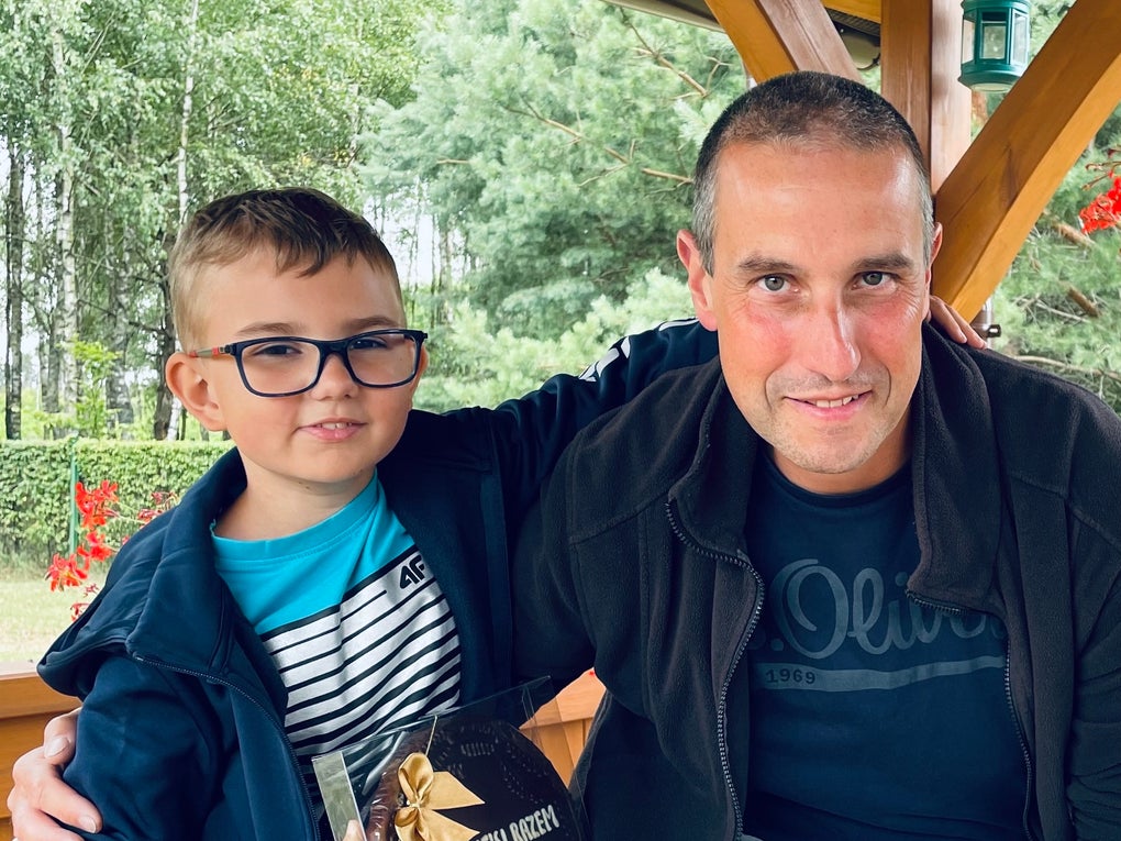 Ein Mann und ein Junge sitzen nebeneinander in einem hölzernen Pavillon. Der Junge hält eine große Schokoladenverpackung mit der Aufschrift „DANKE“ und trägt eine Brille, ein blaues Oberteil und ein gestreiftes Shirt. Der Mann trägt eine dunkle Jacke und ein T-Shirt. Beide blicken in die Kamera. Im Hintergrund sind Bäume und grüne Büsche zu sehen. Ein Mann und ein Junge sitzen nebeneinander in einem hölzernen Pavillon. Der Junge hält eine große Schokoladenverpackung mit der Aufschrift „DANKE“ und trägt eine Brille, ein blaues Oberteil und ein gestreiftes Shirt. Der Mann trägt eine dunkle Jacke und ein T-Shirt. Beide blicken in die Kamera. Im Hintergrund sind Bäume und grüne Büsche zu sehen.