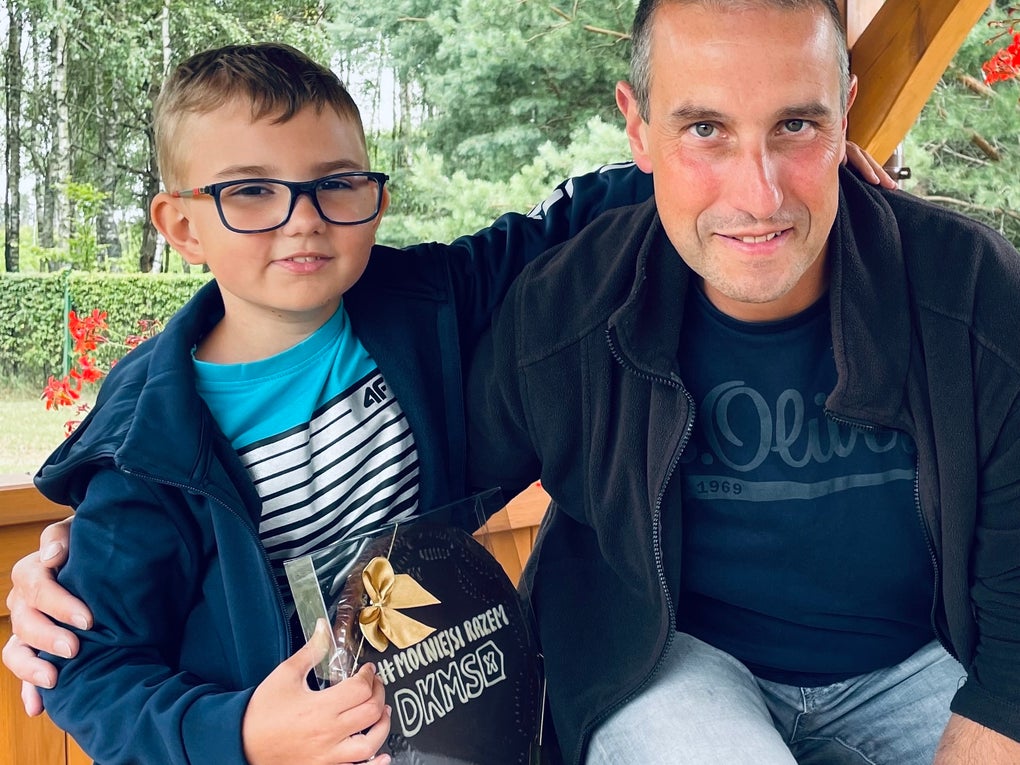 Ein Mann und ein Junge sitzen nebeneinander in einem hölzernen Pavillon. Der Junge hält eine große Schokoladenverpackung mit der Aufschrift „DANKE“ und trägt eine Brille, ein blaues Oberteil und ein gestreiftes Shirt. Der Mann trägt eine dunkle Jacke und ein T-Shirt. Beide blicken in die Kamera. Im Hintergrund sind Bäume und grüne Büsche zu sehen. Ein Mann und ein Junge sitzen nebeneinander in einem hölzernen Pavillon. Der Junge hält eine große Schokoladenverpackung mit der Aufschrift „DANKE“ und trägt eine Brille, ein blaues Oberteil und ein gestreiftes Shirt. Der Mann trägt eine dunkle Jacke und ein T-Shirt. Beide blicken in die Kamera. Im Hintergrund sind Bäume und grüne Büsche zu sehen.