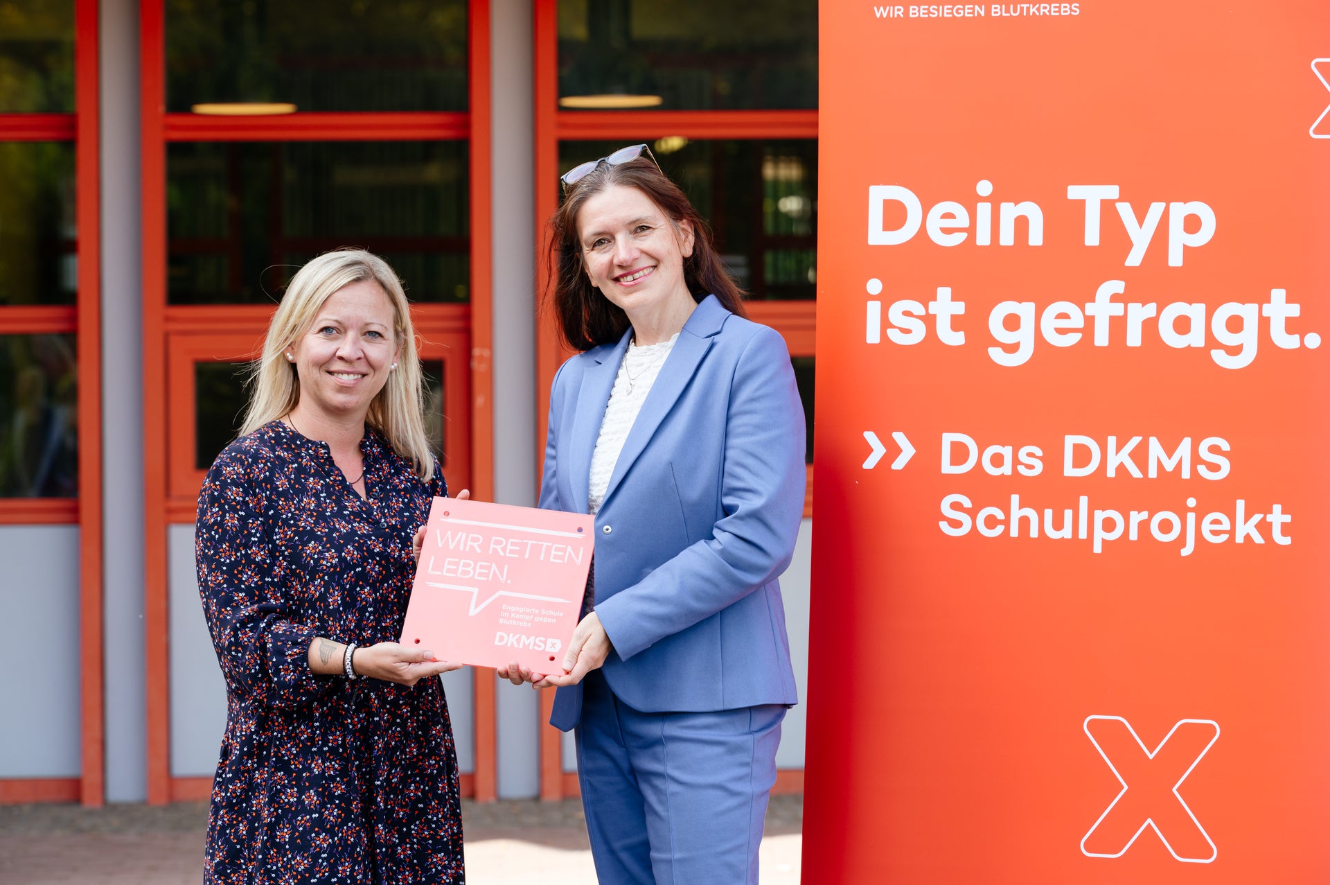 Zwei Frauen posieren für ein Foto, daneben ein Banner mit der Aufschrift: "Dein Typ ist gefragt. Das DKMS Schulprojekt" Zwei Frauen posieren für ein Foto, daneben ein Banner mit der Aufschrift: "Dein Typ ist gefragt. Das DKMS Schulprojekt"