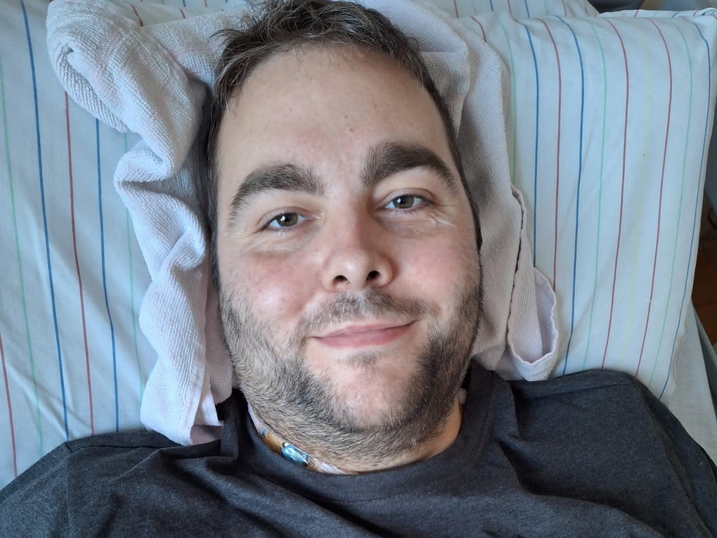 Ein Mann liegt auf einem gestreiften Kopfkissen und blickt in die Kamera. Er trägt ein dunkles T-Shirt, im Halsbereich ist ein medizinischer Zugang sichtbar. Unter seinem Kopf liegt zusätzlich ein weißes Handtuch als Stütze. Ein Mann liegt auf einem gestreiften Kopfkissen und blickt in die Kamera. Er trägt ein dunkles T-Shirt, im Halsbereich ist ein medizinischer Zugang sichtbar. Unter seinem Kopf liegt zusätzlich ein weißes Handtuch als Stütze.