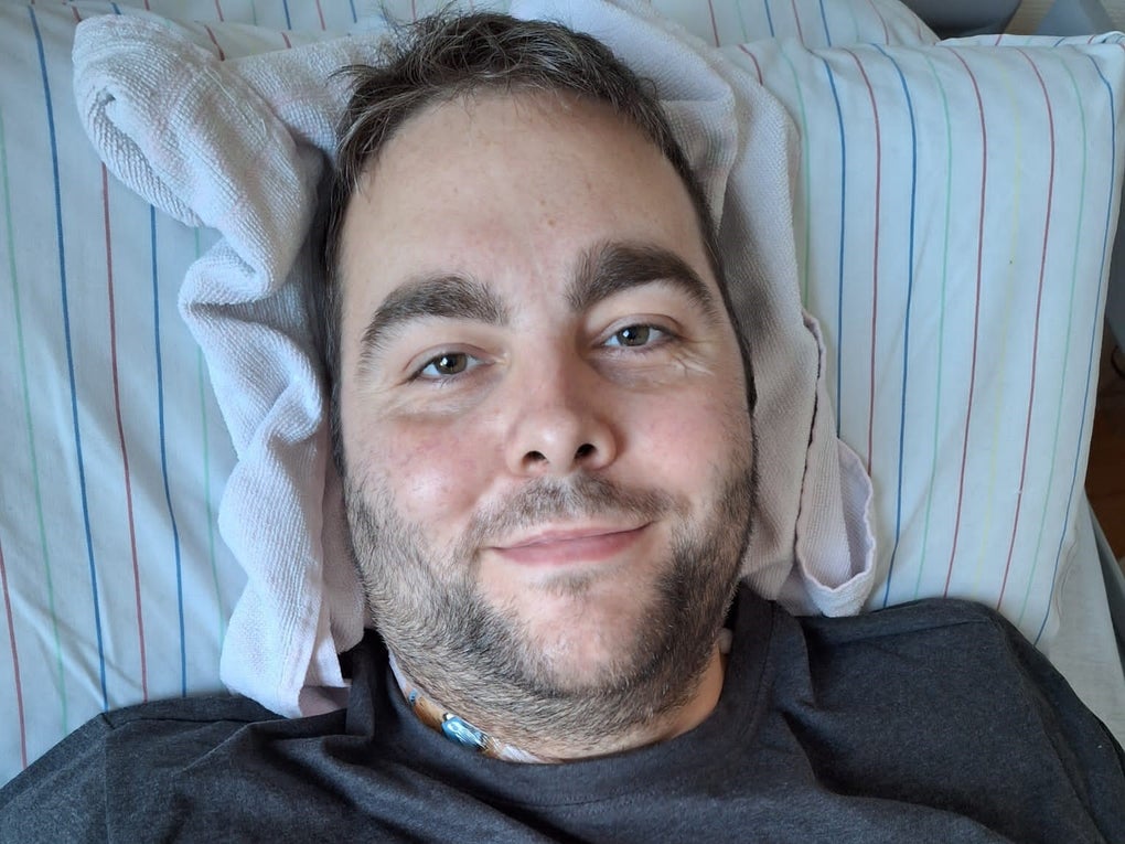 Ein Mann liegt auf einem gestreiften Kopfkissen und blickt in die Kamera. Er trägt ein dunkles T-Shirt, im Halsbereich ist ein medizinischer Zugang sichtbar. Unter seinem Kopf liegt zusätzlich ein weißes Handtuch als Stütze. Ein Mann liegt auf einem gestreiften Kopfkissen und blickt in die Kamera. Er trägt ein dunkles T-Shirt, im Halsbereich ist ein medizinischer Zugang sichtbar. Unter seinem Kopf liegt zusätzlich ein weißes Handtuch als Stütze.