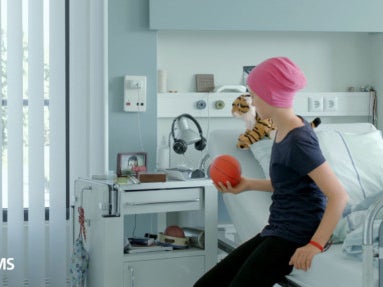 Screenshot aus einem Werbespot. Rinah sitzt auf einem Bett eines Krankenhauszimmers. Links unten im Bild der Text vox.de/dkms Screenshot aus einem Werbespot. Rinah sitzt auf einem Bett eines Krankenhauszimmers. Links unten im Bild der Text vox.de/dkms