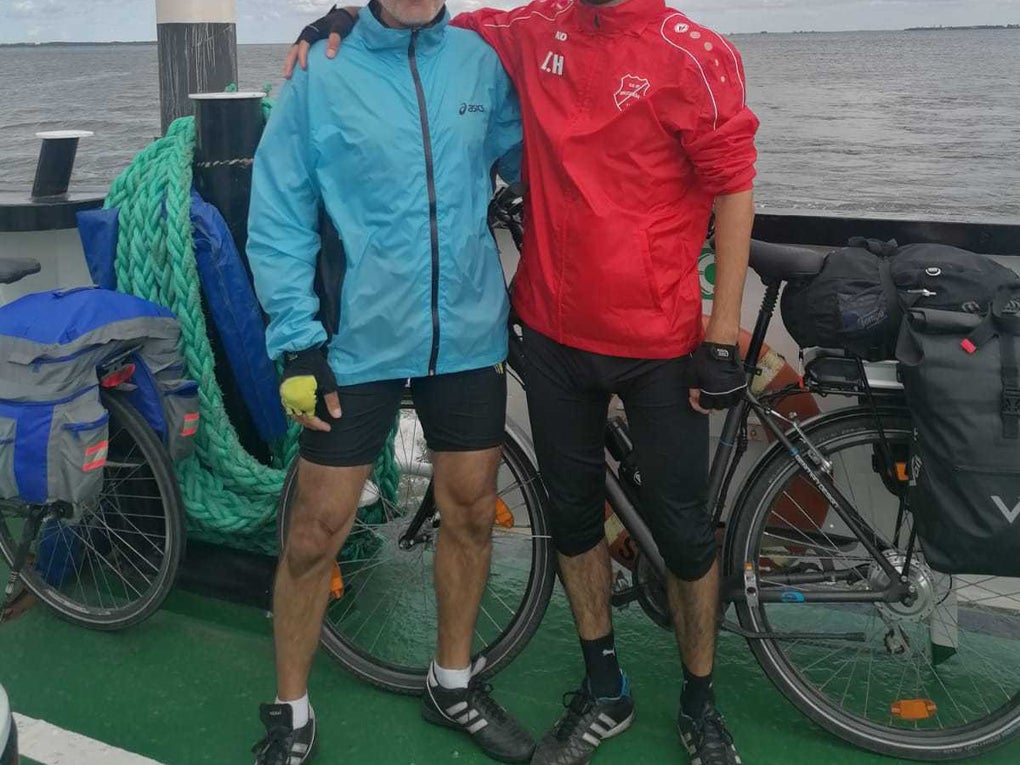 Zwei Männer stehen mit Fahrradbekleidung auf einer Fähre am Wasser. Der ältere Mann links trägt eine blaue Jacke und einen Fahrradhelm, der jüngere Mann rechts eine rote Sportjacke und Sonnenbrille. Beide haben ihre Fahrräder dabei, die mit Gepäcktaschen beladen sind. Im Hintergrund ist eine weite Wasserfläche unter einem teils bewölkten Himmel zu sehen. Zwei Männer stehen mit Fahrradbekleidung auf einer Fähre am Wasser. Der ältere Mann links trägt eine blaue Jacke und einen Fahrradhelm, der jüngere Mann rechts eine rote Sportjacke und Sonnenbrille. Beide haben ihre Fahrräder dabei, die mit Gepäcktaschen beladen sind. Im Hintergrund ist eine weite Wasserfläche unter einem teils bewölkten Himmel zu sehen.