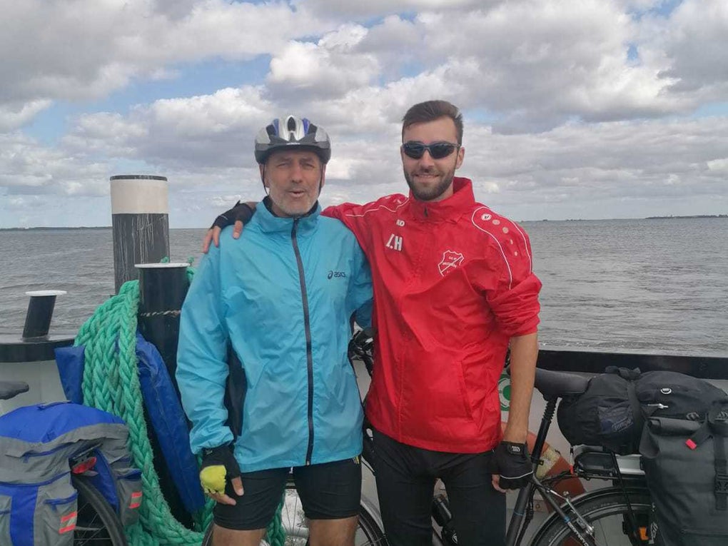 Zwei Männer stehen mit Fahrradbekleidung auf einer Fähre am Wasser. Der ältere Mann links trägt eine blaue Jacke und einen Fahrradhelm, der jüngere Mann rechts eine rote Sportjacke und Sonnenbrille. Beide haben ihre Fahrräder dabei, die mit Gepäcktaschen beladen sind. Im Hintergrund ist eine weite Wasserfläche unter einem teils bewölkten Himmel zu sehen. Zwei Männer stehen mit Fahrradbekleidung auf einer Fähre am Wasser. Der ältere Mann links trägt eine blaue Jacke und einen Fahrradhelm, der jüngere Mann rechts eine rote Sportjacke und Sonnenbrille. Beide haben ihre Fahrräder dabei, die mit Gepäcktaschen beladen sind. Im Hintergrund ist eine weite Wasserfläche unter einem teils bewölkten Himmel zu sehen.