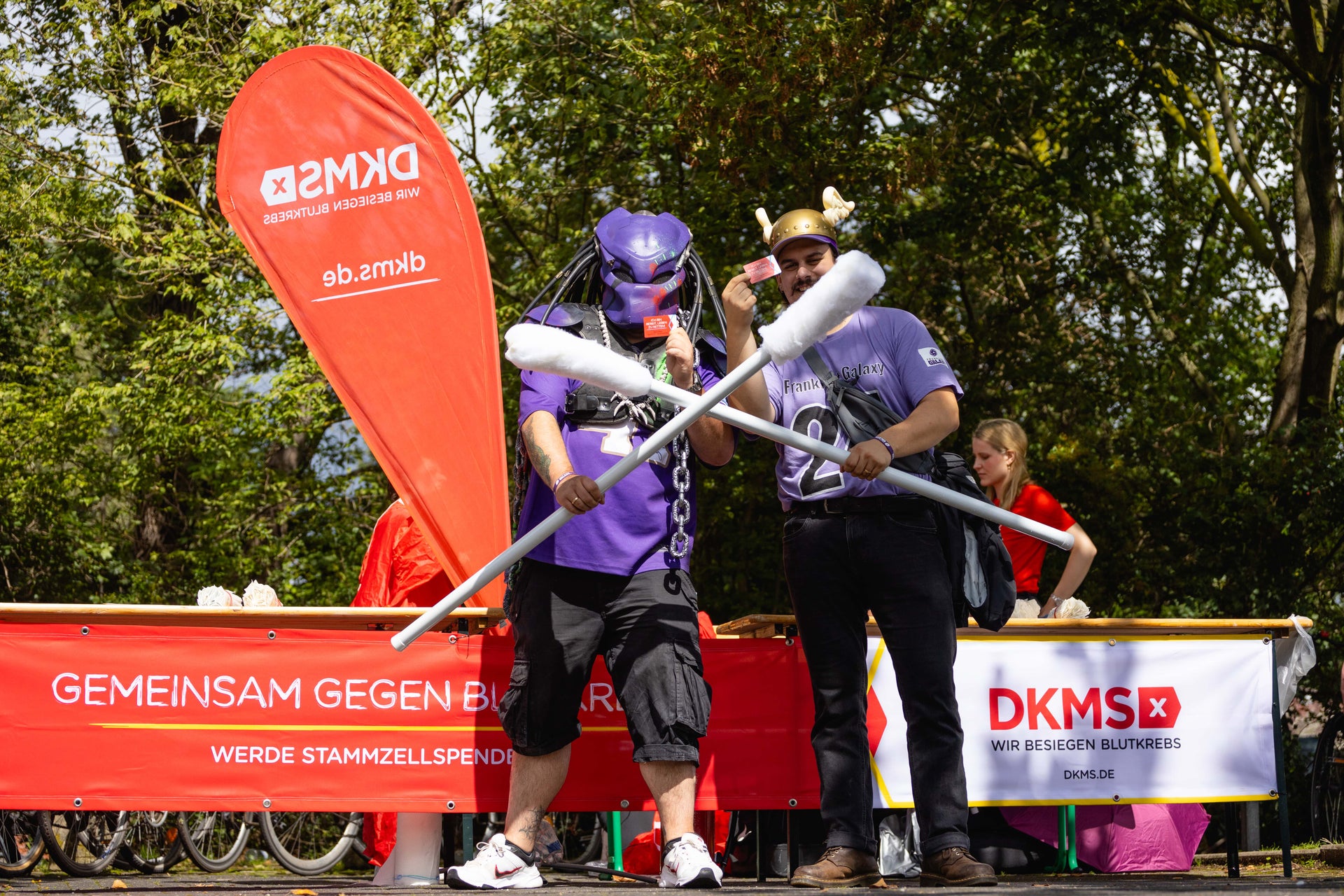 Zwei Personen stehen vor einem DKMS-Aktionsstand im Freien. Beide tragen Fan- oder Kostümkleidung, darunter ein Wikingerhelm und ein Helm mit Maske. Sie halten zwei übergroße Wattestäbchen gekreuzt in den Händen. Im Hintergrund sind ein rotes DKMS-Banner mit der Aufschrift „Gemeinsam gegen Blutkrebs – Werde Stammzellspender:in“ und ein Infostand mit weiteren Personen zu sehen. Bäume und Fahrräder befinden sich im Hintergrund. Zwei Personen stehen vor einem DKMS-Aktionsstand im Freien. Beide tragen Fan- oder Kostümkleidung, darunter ein Wikingerhelm und ein Helm mit Maske. Sie halten zwei übergroße Wattestäbchen gekreuzt in den Händen. Im Hintergrund sind ein rotes DKMS-Banner mit der Aufschrift „Gemeinsam gegen Blutkrebs – Werde Stammzellspender:in“ und ein Infostand mit weiteren Personen zu sehen. Bäume und Fahrräder befinden sich im Hintergrund.