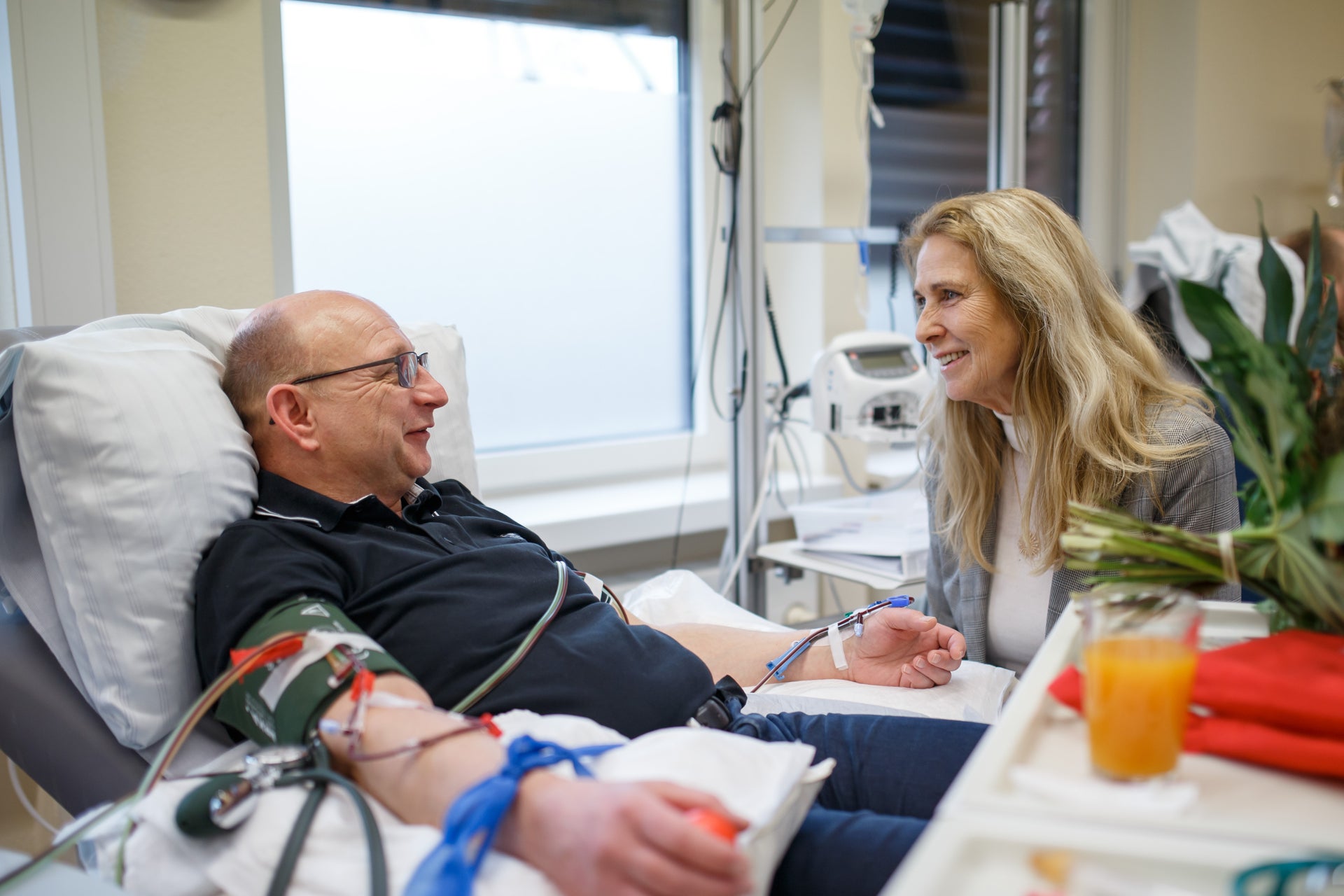 Ein Mann liegt in einem Klinikbett und spendet Blutstammzellen. Er trägt eine Blutdruckmanschette am Arm, mehrere Schläuche sind angeschlossen. Neben ihm sitzt eine Frau mit langen blonden Haaren, die ihn freundlich anlächelt. Auf dem Tisch vor ihnen stehen ein Glas Orangensaft, ein Blumenstrauß und rote Stoffservietten.
Ein Mann liegt in einem Klinikbett und spendet Blutstammzellen. Er trägt eine Blutdruckmanschette am Arm, mehrere Schläuche sind angeschlossen. Neben ihm sitzt eine Frau mit langen blonden Haaren, die ihn freundlich anlächelt. Auf dem Tisch vor ihnen stehen ein Glas Orangensaft, ein Blumenstrauß und rote Stoffservietten.