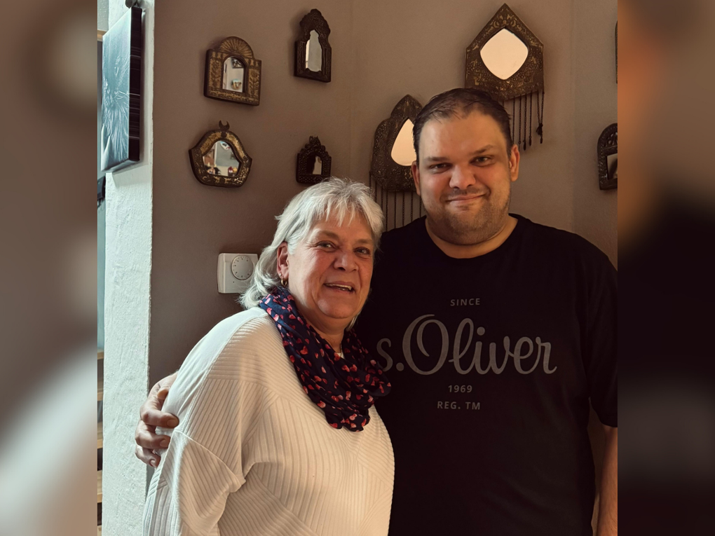 Ein Mann und eine Frau stehen Arm in Arm vor einer braun gestrichenen Wand und lächeln in die Kamera. Ein Mann und eine Frau stehen Arm in Arm vor einer braun gestrichenen Wand und lächeln in die Kamera.