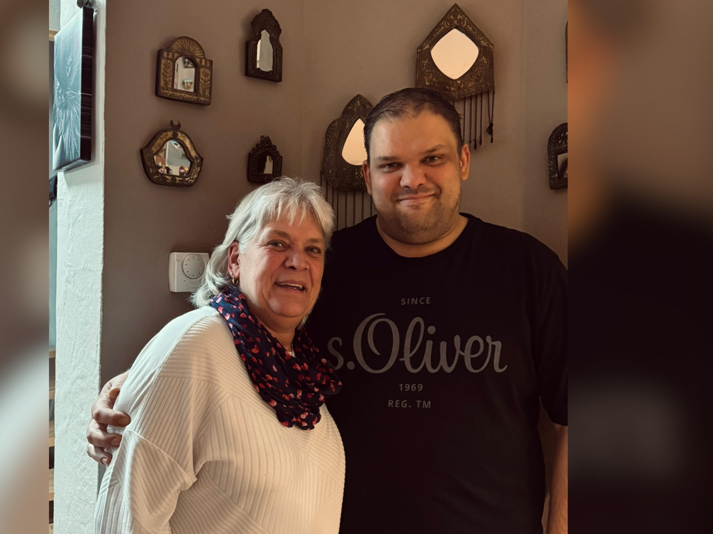 Ein Mann und eine Frau stehen Arm in Arm vor einer braun gestrichenen Wand und lächeln in die Kamera. Ein Mann und eine Frau stehen Arm in Arm vor einer braun gestrichenen Wand und lächeln in die Kamera.