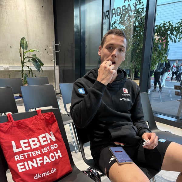 Ein Mann, der auf einer schwarzen Stuhlreihe sitzt. Neben ihm liegt ein roter DKMS Beutel. Ein Mann, der auf einer schwarzen Stuhlreihe sitzt. Neben ihm liegt ein roter DKMS Beutel.