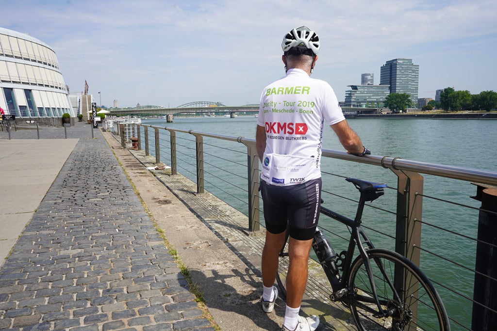 Ein Mann, der einen Helm trägt und ein Fahrrad hält, das an einem Geländer lehnt. Im Hintergrund ist ein Gewässer. Ein Mann, der einen Helm trägt und ein Fahrrad hält, das an einem Geländer lehnt. Im Hintergrund ist ein Gewässer.