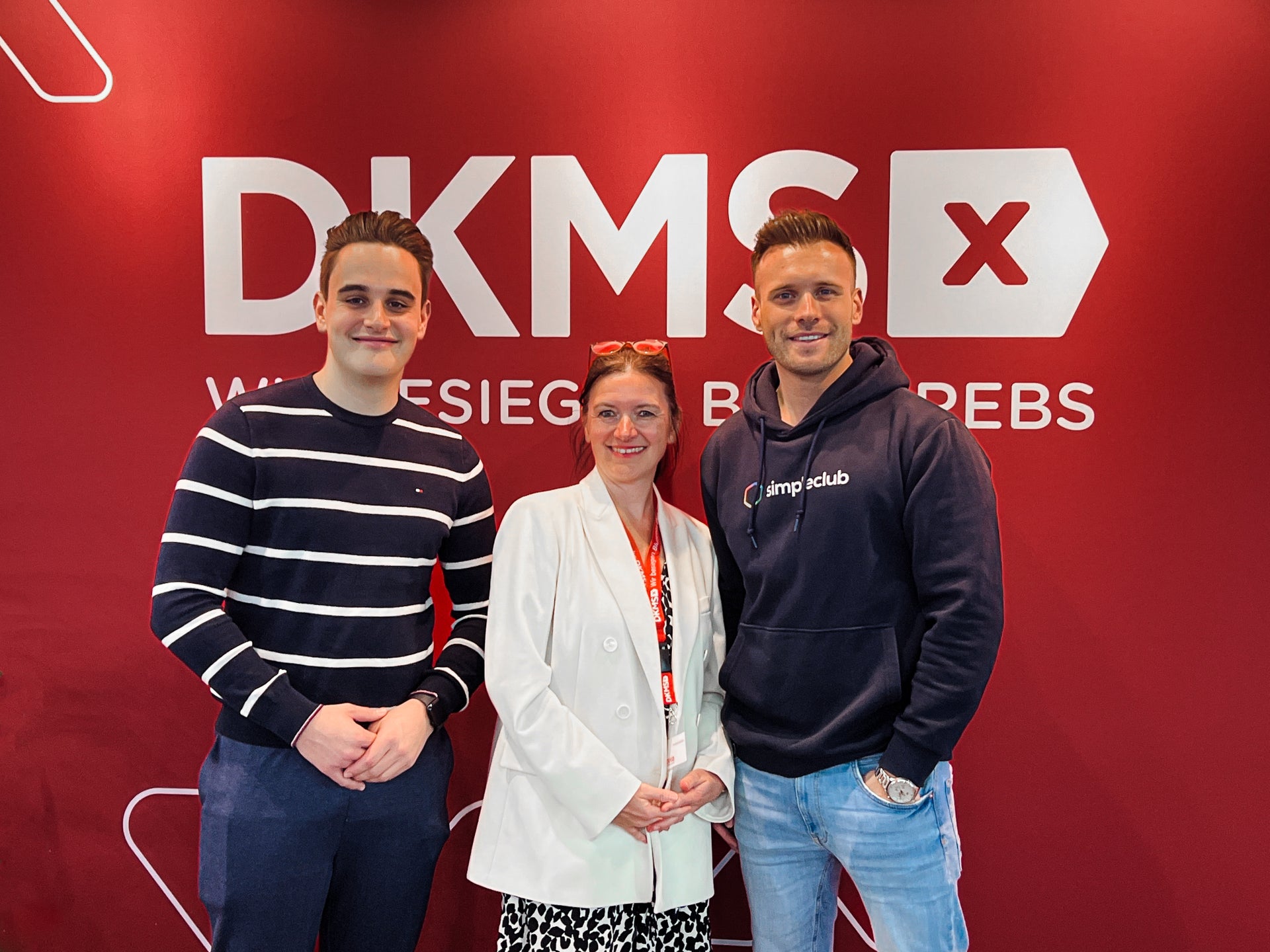 Drei Personen stehen lächelnd vor einer roten DKMS-Wand mit dem Schriftzug "DKMS – Wir besiegen Blutkrebs". In der Mitte steht eine Frau in weißem Blazer, links ein junger Mann im dunkel gestreiften Pullover, rechts ein Mann in einem dunkelblauen simpleclub-Hoodie. Drei Personen stehen lächelnd vor einer roten DKMS-Wand mit dem Schriftzug "DKMS – Wir besiegen Blutkrebs". In der Mitte steht eine Frau in weißem Blazer, links ein junger Mann im dunkel gestreiften Pullover, rechts ein Mann in einem dunkelblauen simpleclub-Hoodie.