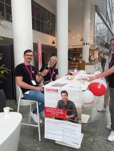 Zwei Personen mit pinken DKMS-Lanyards sitzen an einem DKMS-Informationsstand in einem modernen, hellen Gebäude mit hoher Glasfassade. Beide halten Wattestäbchen in der Hand und lächeln in die Kamera. Auf dem Tisch liegen Registrierungsunterlagen und DKMS-Materialien, darunter rote Luftballons. Im Vordergrund hängt ein Plakat mit dem Slogan „Das beste Mittel gegen Blutkrebs bist du“ und dem Aufruf zur Stammzellspende. Eine dritte Person steht am rechten Bildrand, teilweise im Anschnitt. Zwei Personen mit pinken DKMS-Lanyards sitzen an einem DKMS-Informationsstand in einem modernen, hellen Gebäude mit hoher Glasfassade. Beide halten Wattestäbchen in der Hand und lächeln in die Kamera. Auf dem Tisch liegen Registrierungsunterlagen und DKMS-Materialien, darunter rote Luftballons. Im Vordergrund hängt ein Plakat mit dem Slogan „Das beste Mittel gegen Blutkrebs bist du“ und dem Aufruf zur Stammzellspende. Eine dritte Person steht am rechten Bildrand, teilweise im Anschnitt.