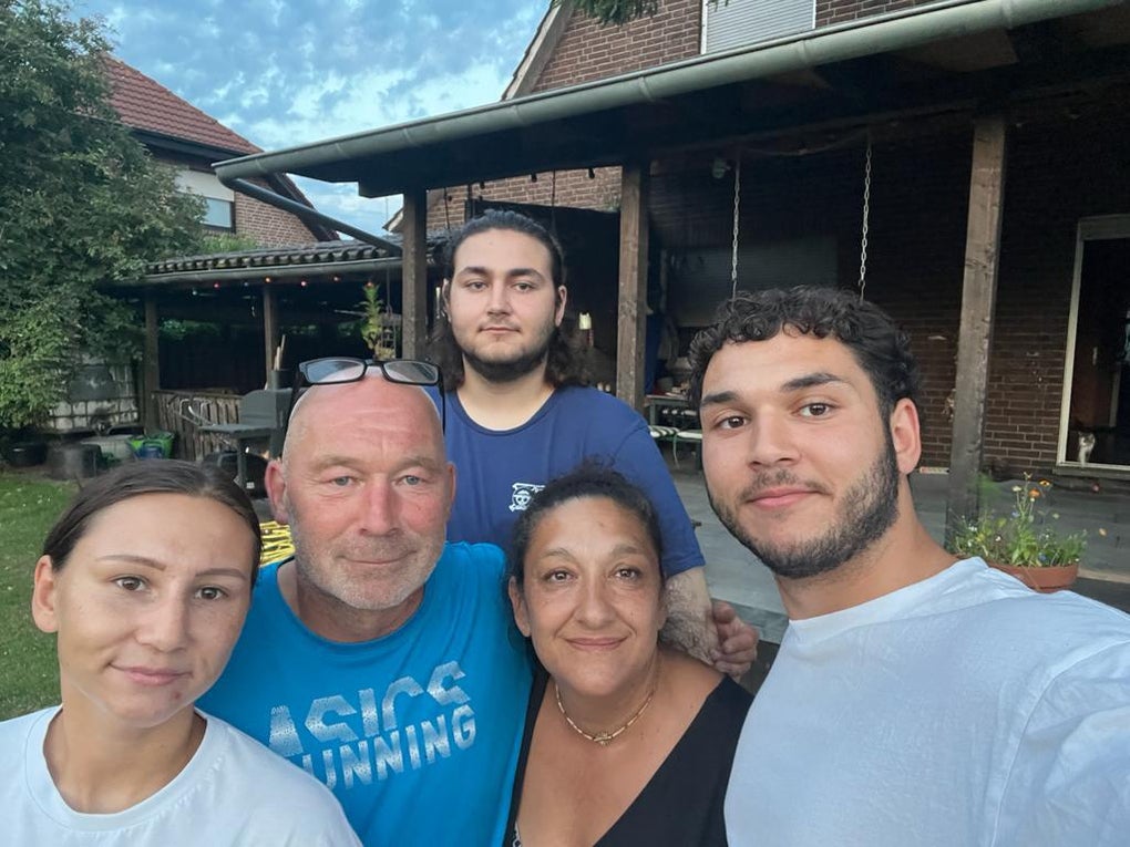 Fünf Personen stehen eng beieinander und lächeln in die Kamera. Im Hintergrund ist ein Backsteinhaus mit überdachter Terrasse und Garten zu sehen. Der Himmel ist leicht bewölkt. Fünf Personen stehen eng beieinander und lächeln in die Kamera. Im Hintergrund ist ein Backsteinhaus mit überdachter Terrasse und Garten zu sehen. Der Himmel ist leicht bewölkt.