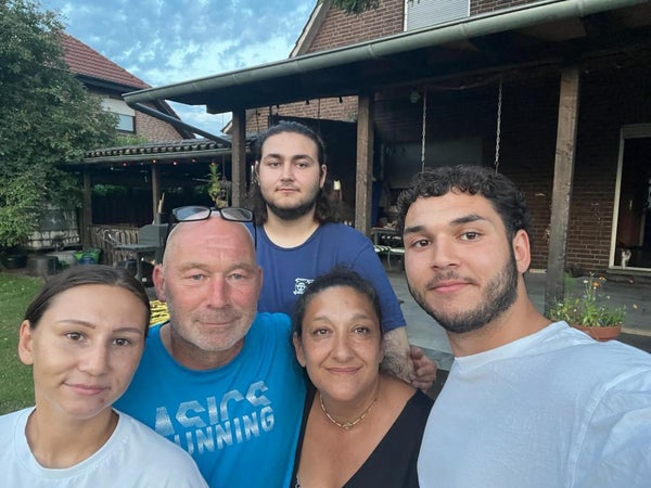 Fünf Personen stehen eng beieinander und lächeln in die Kamera. Im Hintergrund ist ein Backsteinhaus mit überdachter Terrasse und Garten zu sehen. Der Himmel ist leicht bewölkt. Fünf Personen stehen eng beieinander und lächeln in die Kamera. Im Hintergrund ist ein Backsteinhaus mit überdachter Terrasse und Garten zu sehen. Der Himmel ist leicht bewölkt.